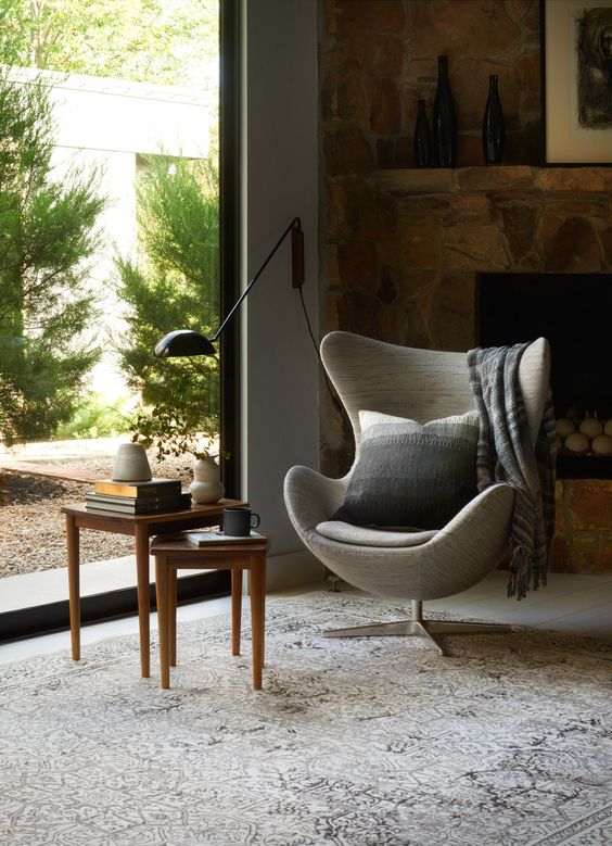 a cozy nook with a fireplace clad with stone, a grey Egg chair with a pillow, a couple of side tables and a gorgeous view