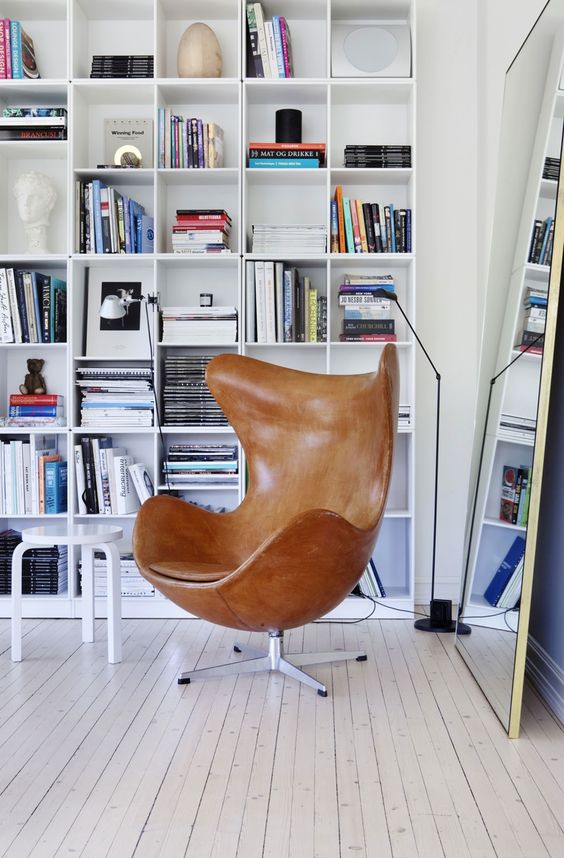 a cozy reading space with a large bookcase, an amber leather Egg chair, a large floor mirror and a black floor lamp