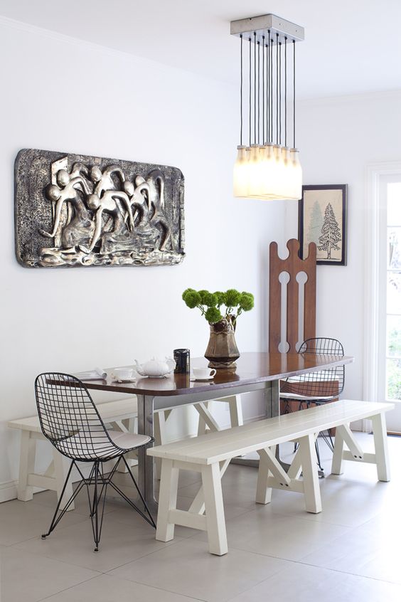 a dining room with a stained table, white benches, black Eames wire chairs, a catchy bottle chandelier and some decor