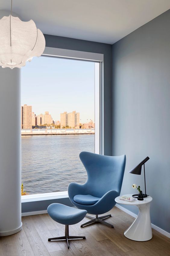 a gorgeous nook with a floor to ceiling window, grey walls, a blue Egg chair and a footrest, a white side table with a black lamp