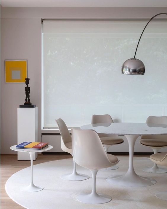 a neutral dining room with a large window, an oval table, neutral Tulip chairs, a round rug, a large lamp and some decor