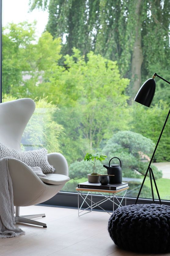a peaceful nook with a glazed wall and a greenery view, a white Egg chair with pillows, a black pouf, a black floor lamp and a side table