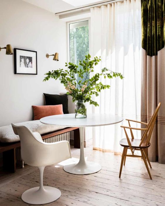 a pretty breakfast nook with a bench over a radiator, a round table, a Tulip chair and a stained one, some pillows and decor