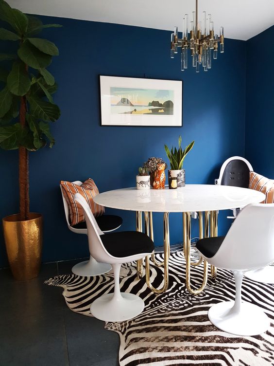 a refined and bold dining room with navy walls, a table with gold piping legs, black Tulip chairs, a zebra print rug, a cool chandelier