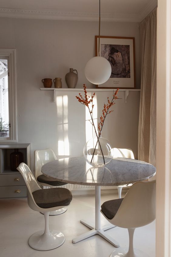 a small neutral dining nook with a grey storage unit, a round table, grey tulip chairs, a shelf with some decor
