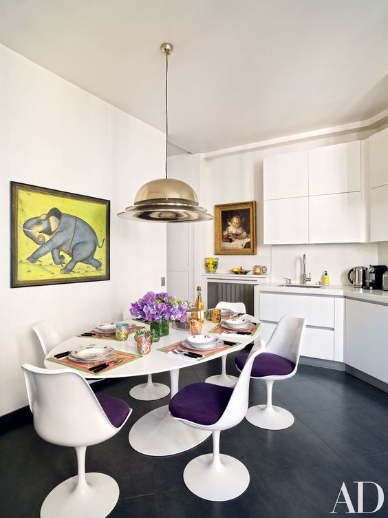 a stylish kitchen with a dining space with sleek white cabinets, an oval table with purple Tulip chairs, a cool pendant lamp and a bold artwork