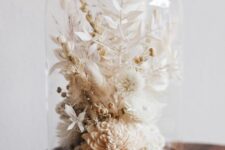 a stylish neutral dried flower arrangement of neutral dried blooms and leaves placed into a cloche with a wooden base