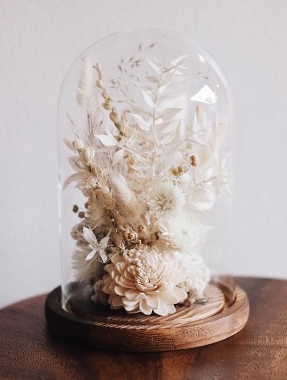 a stylish neutral dried flower arrangement of neutral dried blooms and leaves placed into a cloche with a wooden base