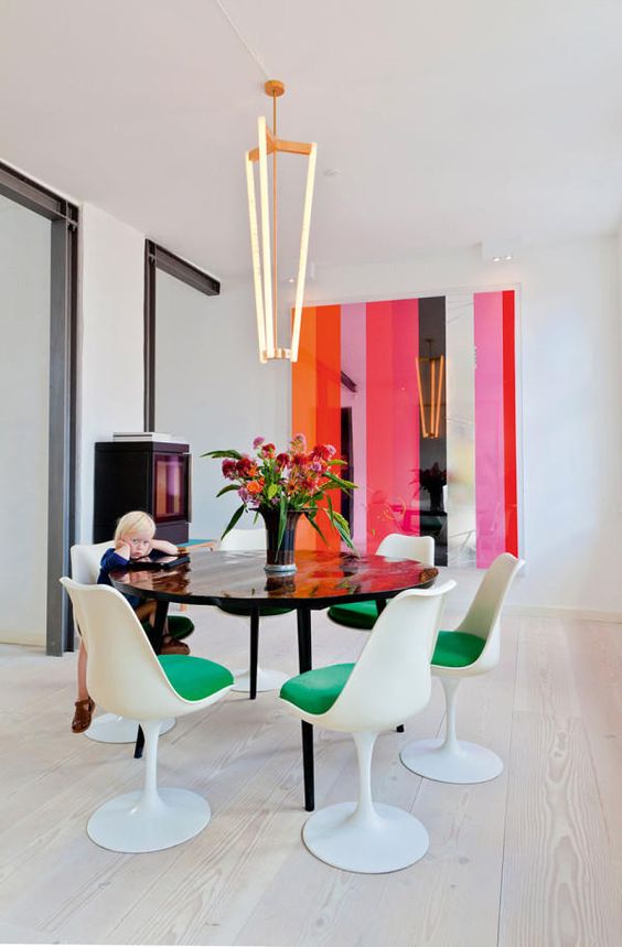 a super bright dining room with bold stripes on the wall, a storage unit, a black table and green Tulip chairs plus a gorgeous modern chandelier