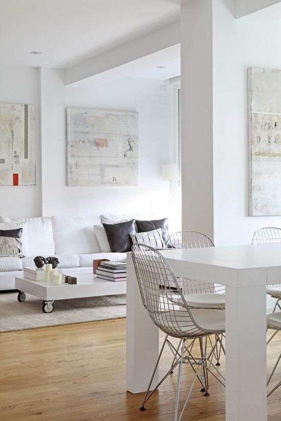 a white dining and living room with a white sofa and dark pillows, a low coffee table on casters, a white dining table and Eames wire chairs