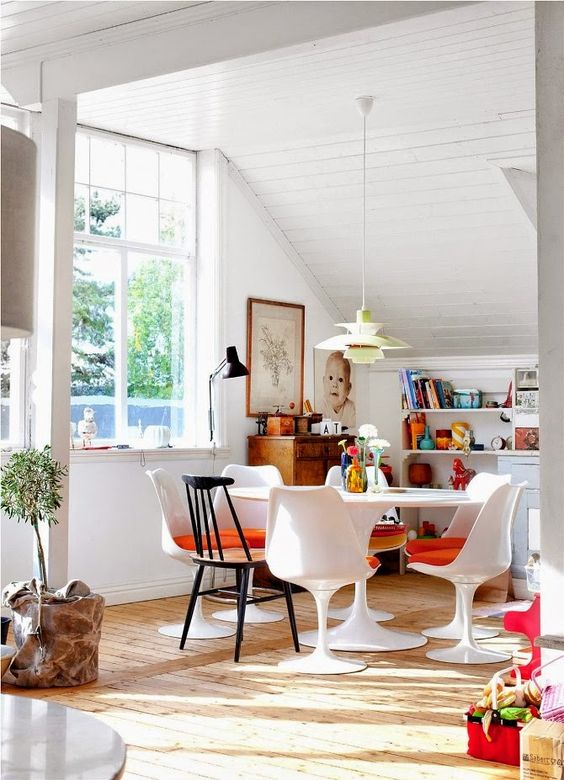 an eclectic dining space with open shelves with books, a vintage sideboard, a round table and orange Tulip chairs, a pendant lamp and a black table one