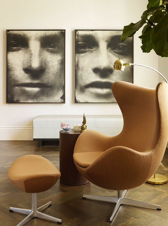 an elegant space with a white upholstered bench, a side table, a rust-colored Egg chair and a footrest, oversized artwork and a floor lamp