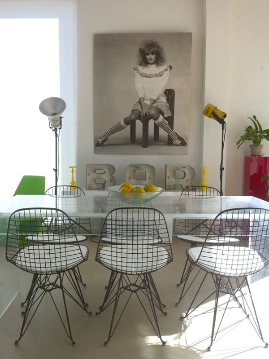 an industrial dining space with a glass table, Eames wire chairs, some industrial lamps and art and a potted plant