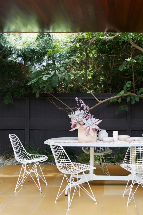 an outdoor dining space with a large white dining table, white Eames wire chairs and some greenery around