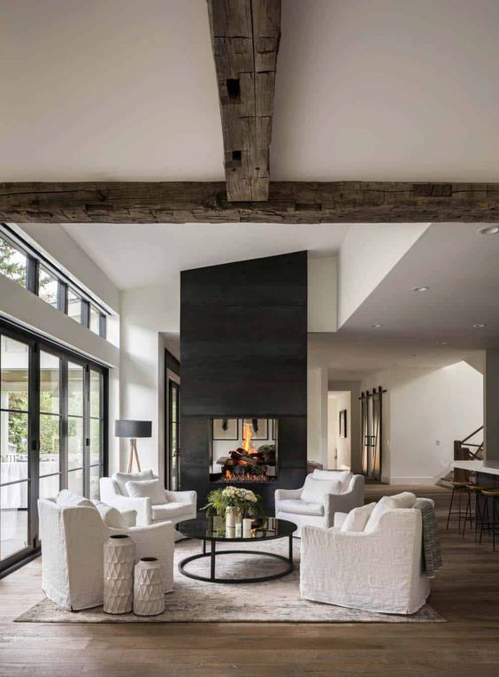 a beautiful modern farmhouse living room with reclaimed wooden beams, a large double-sided fireplace, white seating furniture and a coffee table