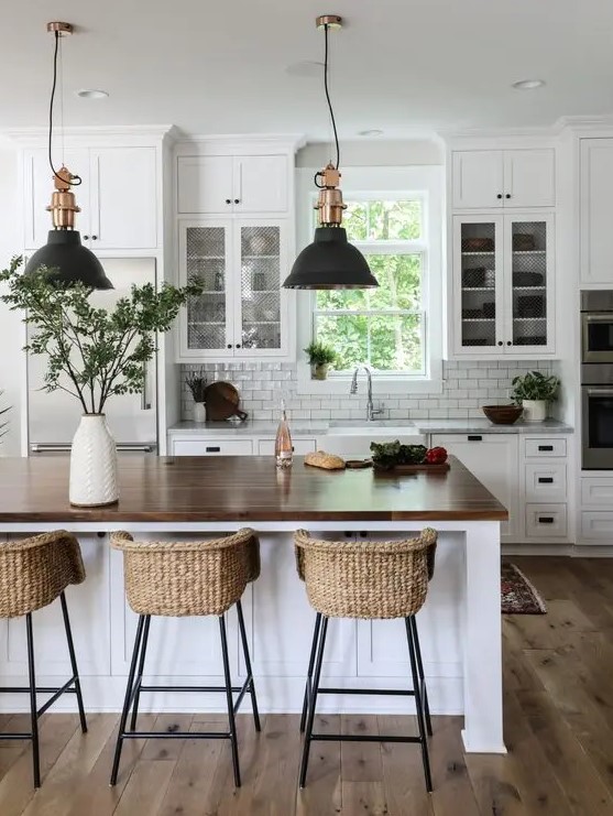 a modern country kitchen in white, with cool cabinets, a large kitchen island with a butcherblock countertop, pendant lamps and woven stools