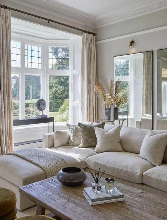 a modern country living room in neutrals, with mirrors, a creamy sectional, a bay window, a wooden table and cool textiles