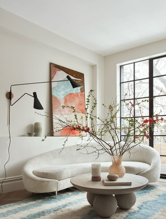 a beautiful living room with a curved creamy sofa, a round coffee table, a bold artwork, black wall lamps and a glazed wall