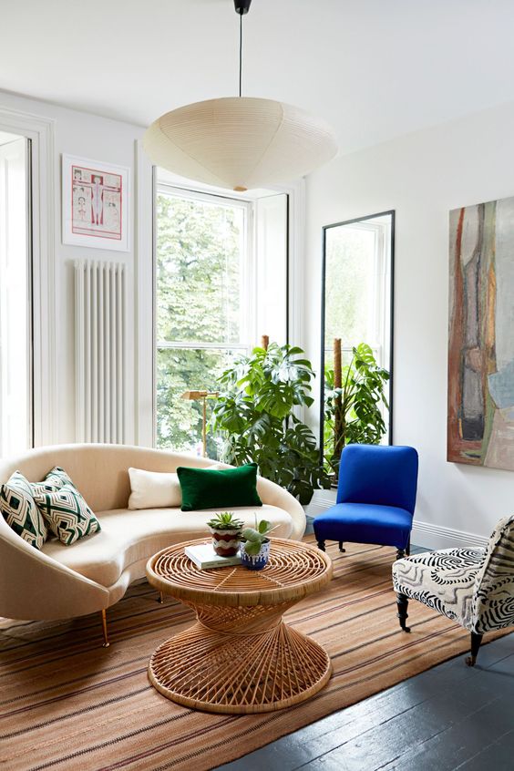 a bright living room with a curved sofa and pillows, a blue and a printed chair, a rattan coffee table, some art and a mirror