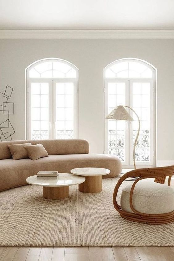 a chic neutral living room with a curved sofa, a duo of coffee tables, a rattan chair, a floor lamp and arched windows