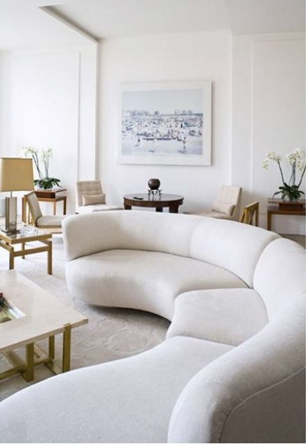 a neutral living room with a white curved sofa, a coffee table, side tables, chairs and an artwork, some potted blooms