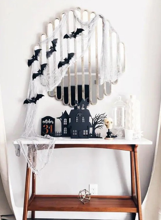 a Halloween console table with cheesecloth, black bats, black houses and a white skull is a bold and cool idea