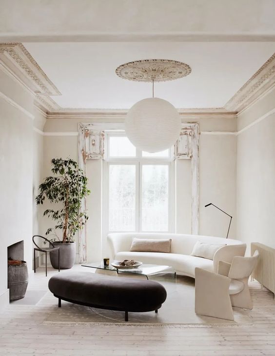 a refined neutral living room with a white curved sofa and a chair, a black curved ottoman, an acrylic coffee table and a potted plant