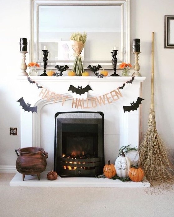 a Halloween mantel with bats, candles, pumpkins, orange blooms, a wheat bundle and some pumpkins and a broom on the floor