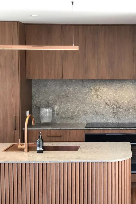 a chic and cozy kitchen with stained cabinets, a fluted curved kitchen island and grey stone countertops and a backsplash