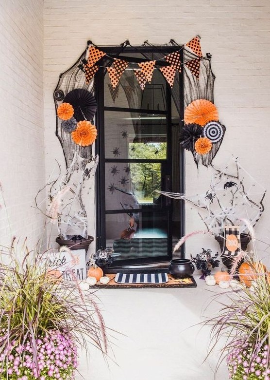 a bold and whimsical Halloween porch with paper fans, buntings, orange and white pumpkins, spiders and branches