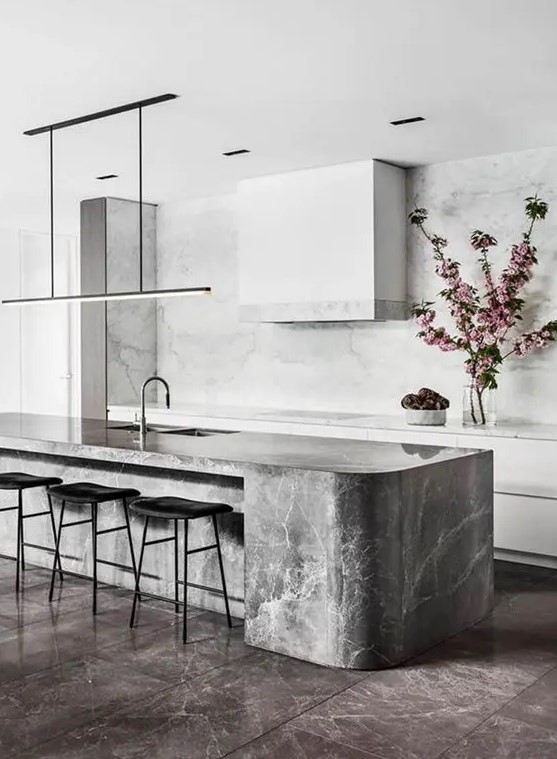 a minimalist white kitchen with a grey curved kitchen island made of a marble slab and a white marble backsplash and countertops