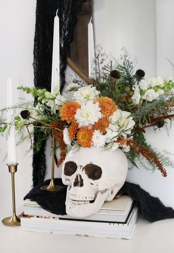 a Halloween decoration of a skull with white and orange blooms, black flowers, greenery and herbs is a nice centerpiece