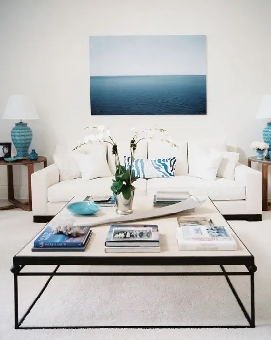 a beahcy living room in cremay and bold blues, a modern coffee table, a large and bold artwork, side tables with blue lamps