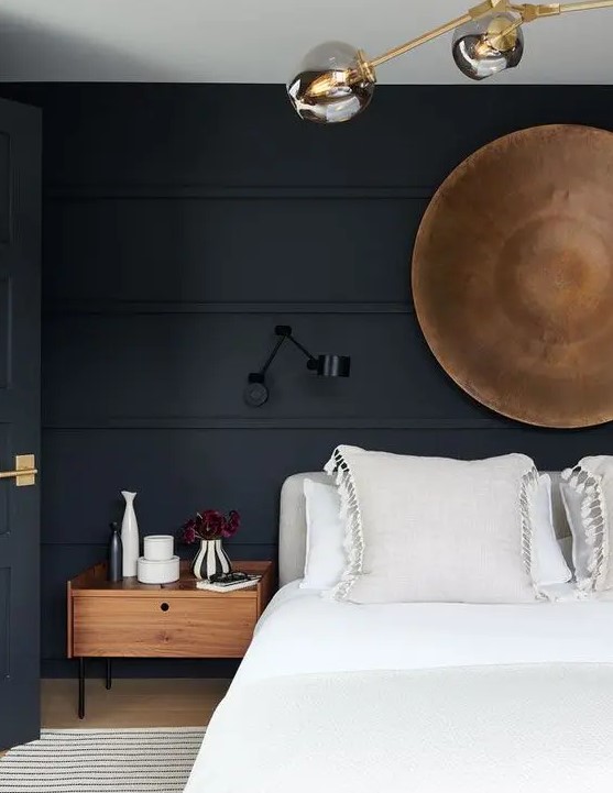 a beautiful moody bedroom with soot walls and a door, a grey upholstered bed with neutral bedding, stained nightstands, some lamps and a wooden bowl on the wall
