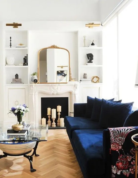 a beautiful neutral living room with a candle fireplace, a navy sofa, built-in shelves, a glass coffee table and lovely gold touches here and there