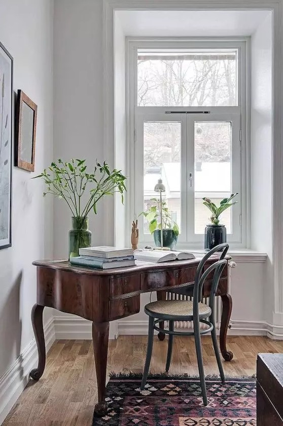 a beautiful vintage home office space with a large heavy stained desk, a grey chair, some potted plants and large books