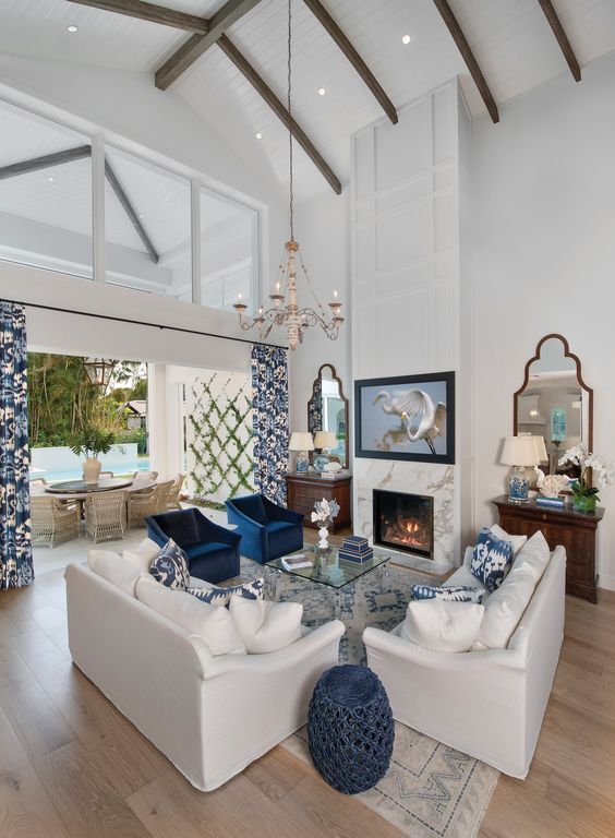 a bold living room with a double-height ceiling, neutral sofas, navy chairs, a glass coffee table, a fireplace and a chandelier