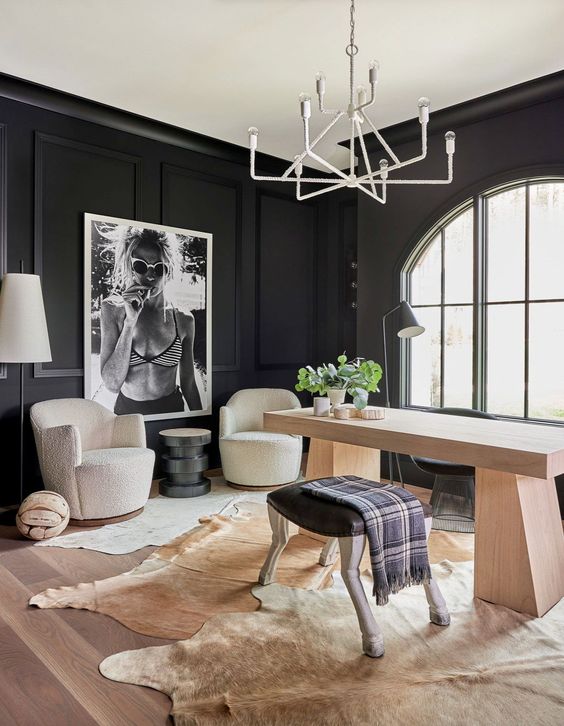 a bold soot home office with trim on the walls, a light-stained desk, a stool, creamy chairs and cowhide rugs