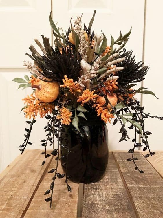 a catchy Halloween flower arrangement of a black jar, greenery, black elements, orange pumpkins and blooms