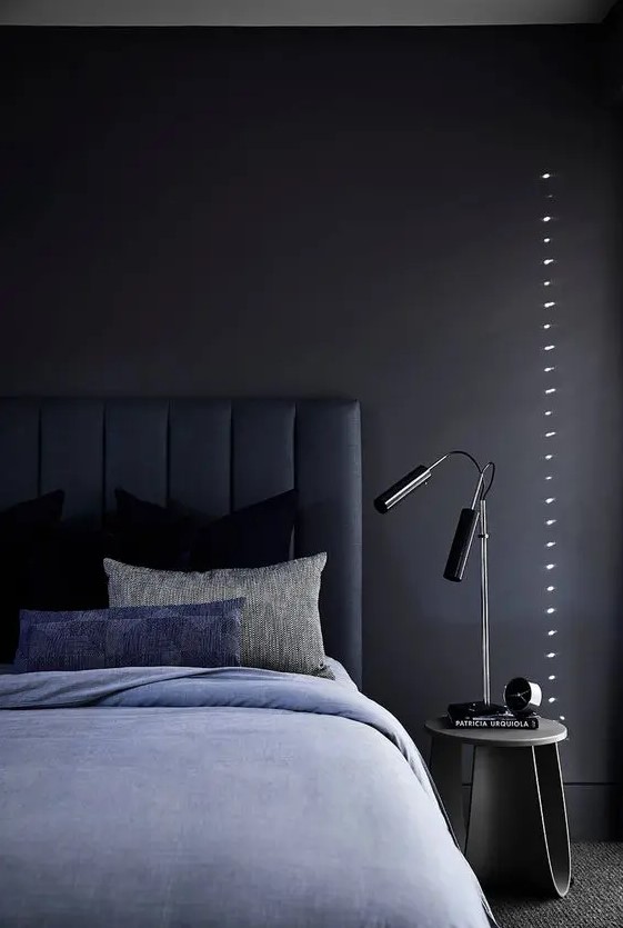 a chic and peaceful moody bedroom with a soot accent wall, a navy upholstered bed and blue bedding, a nightstand, black table lamps