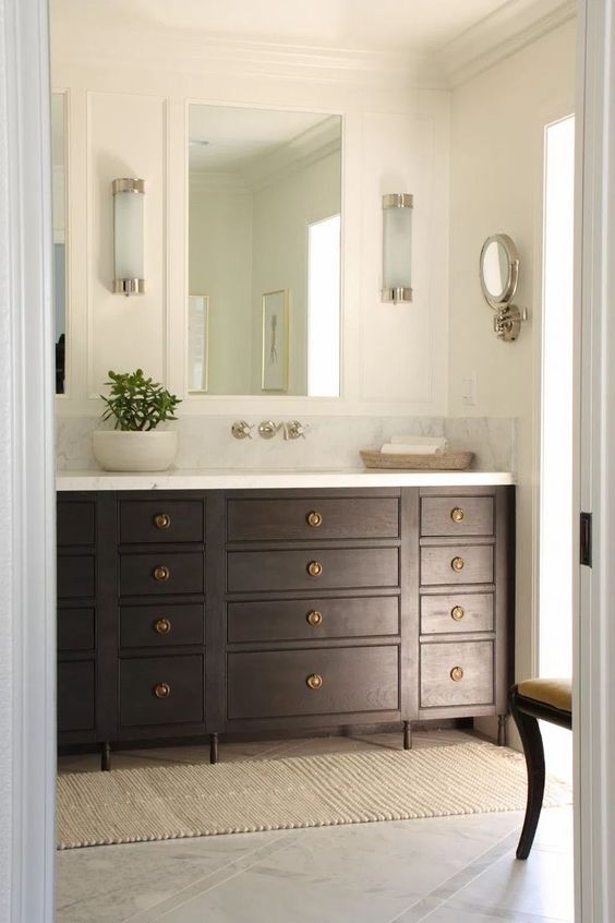 a chic and refined bathroom with a dark-stained vanity, a mirror, some sconces and a vintage chair, a wall mirror