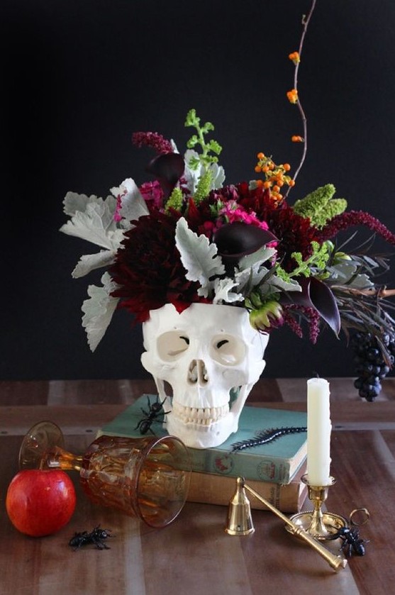 a chic centerpiece of a skull and dark and moody florals, pale and usual greenery and some berries look very cool