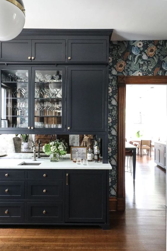 a chic kitchen with soot cabinets, dark floral wallpaper, a mirror backsplash, white stone countertops and shiny knobs
