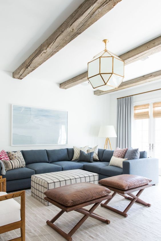 a coastal living room with a blue sectional, a printed ottoman, brown folding stools, a faceted pendant lamp and reclaimed wooden beams