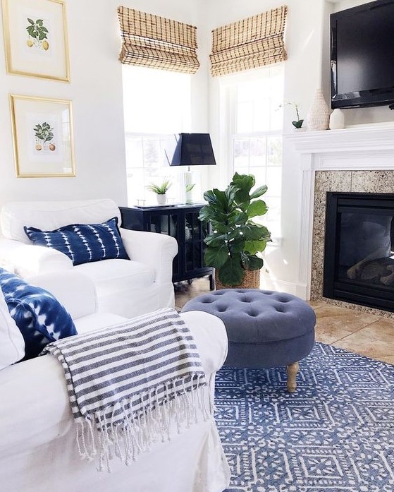 a cozy beach living space with white furniture, a blue ottoman, a printed rug and woven Roman shades