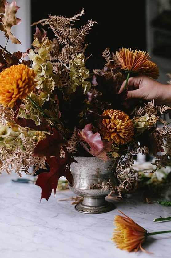 a dark and moody floral centerpiece of a silver urn, rust and mustard blooms, dried leaves and bright fall leaves