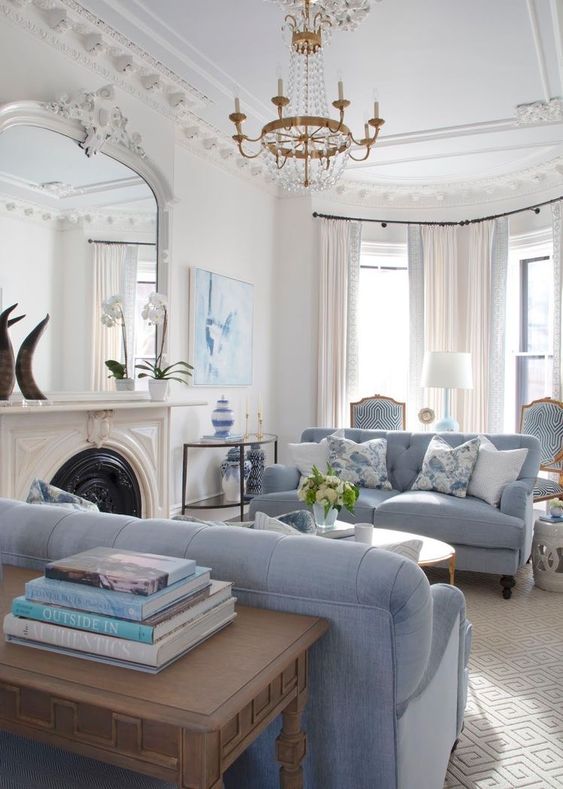 a delicate living room with a French fireplace, a large mirror in an ornated frame, sky blue sofas, side tables and a crystal chandelier