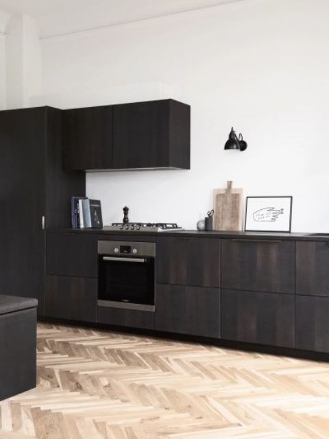 a dramatic dark-stained sleek kitchen with no handles and built-in appliances plus some decor, a black sconce on the wall
