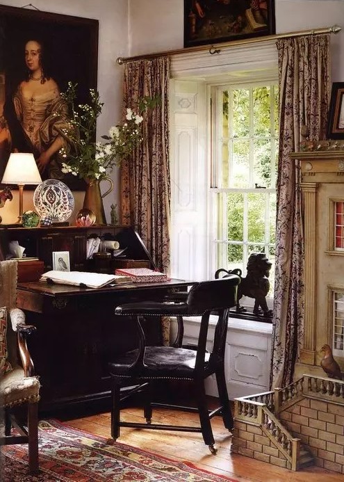 a fab vintage home office with vintage artwork, a dark-stained bureau desk and a black chair, printed curtains and a rug plus some blooms