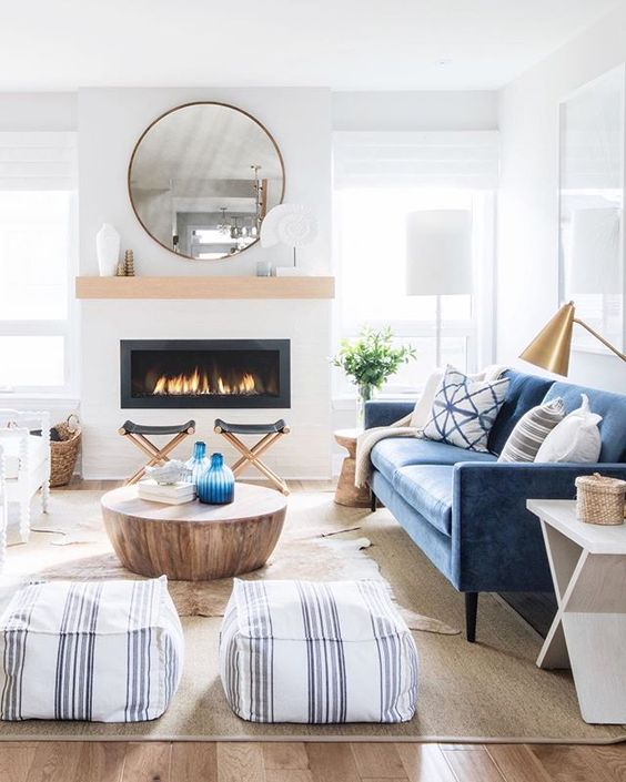 a light-filled coastal living room with a built-in fireplace, a blue sofa, a coffee table, printed poufs, folding stools and a gold lamp